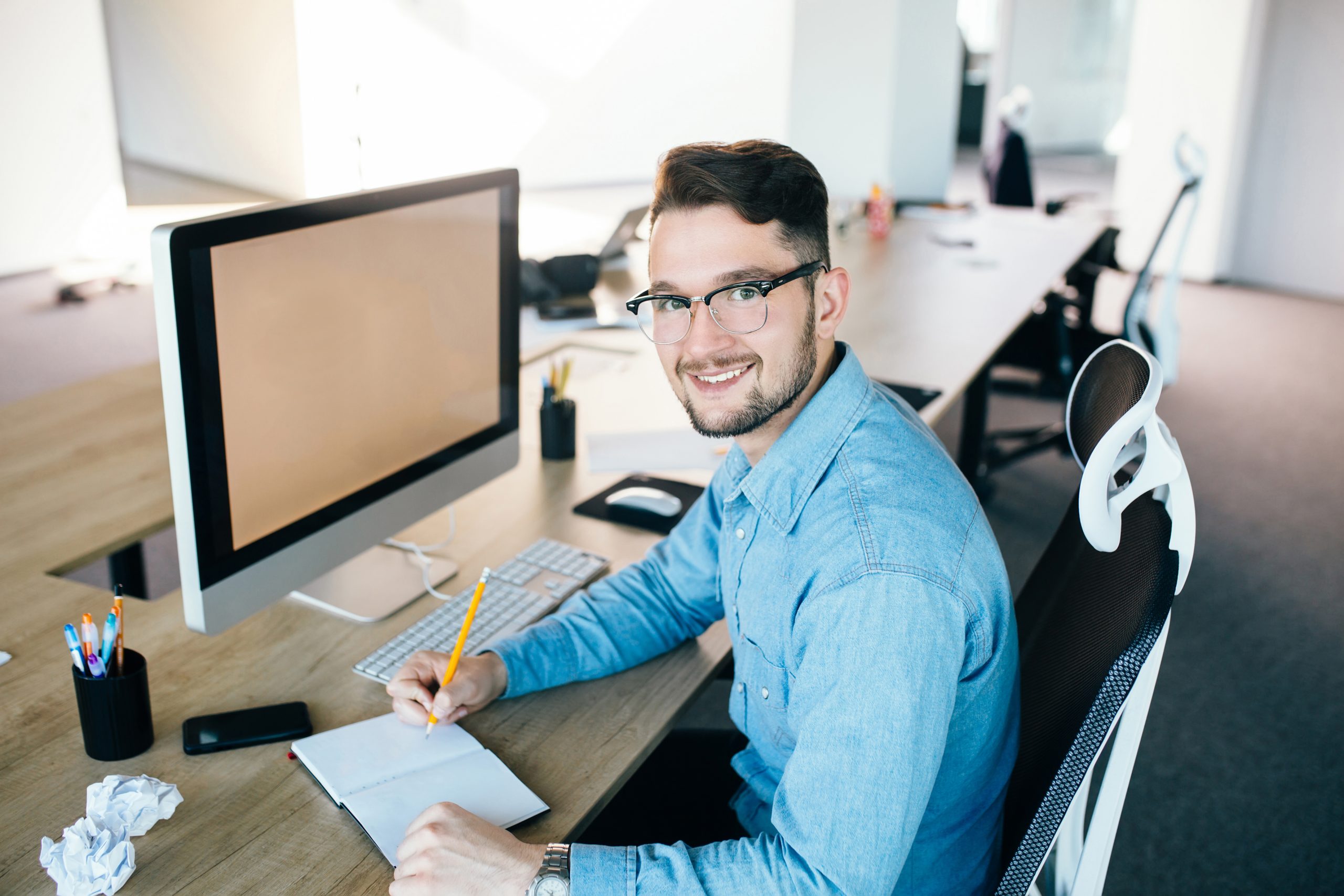 Young man in glassess is working at his workplace in office. He wears blue shirt. He is writing in notebook and smiling to the camera